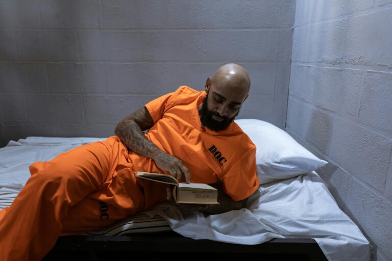 Tattooed inmate reads a book while lying on a bed in a jail cell.